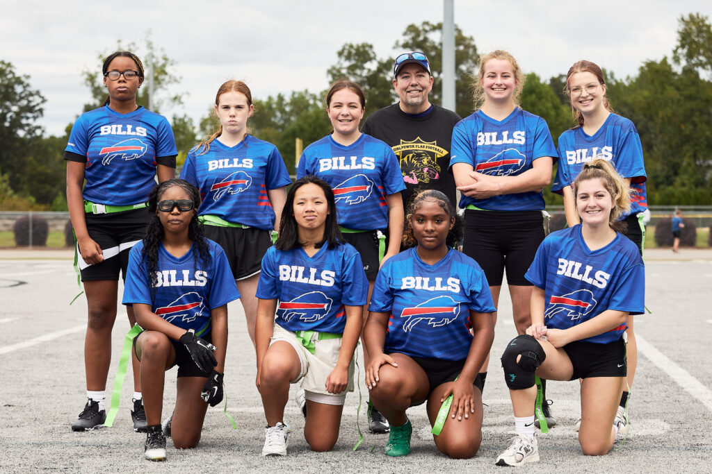 Mooresville girls flag football team posing together in jerseys and flags before game.